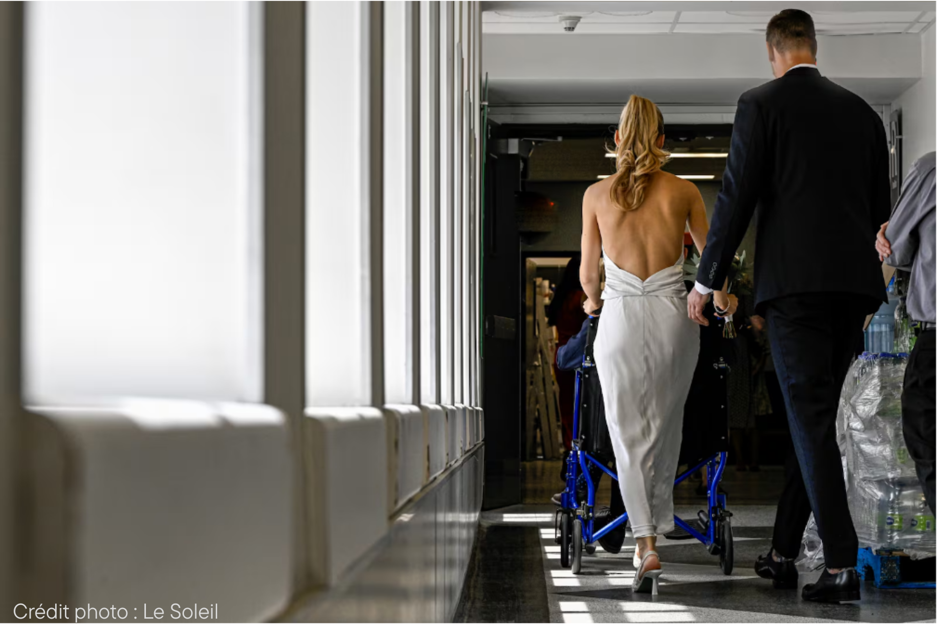 Vue de dos: une mariée pousse un fauteuil roulant dans un corridor d'hôpital